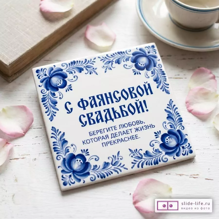 Белая плитка с синим цветочным узором и текстом «С ФАЯНСОВОЙ СВАДЬБОЙ! БЕРЕГИТЕ ЛЮБОВЬ, КОТОРАЯ ДЕЛАЕТ ЖИЗНЬ ПРЕКРАСНЕЕ.», белый фон, розовые лепестки, чашка и книга.